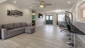 Ein geräumiges Wohnzimmer mit grauem Ecksofa, einem kleinen Hocker, einem Deckenventilator und einem Hängekorbstuhl.