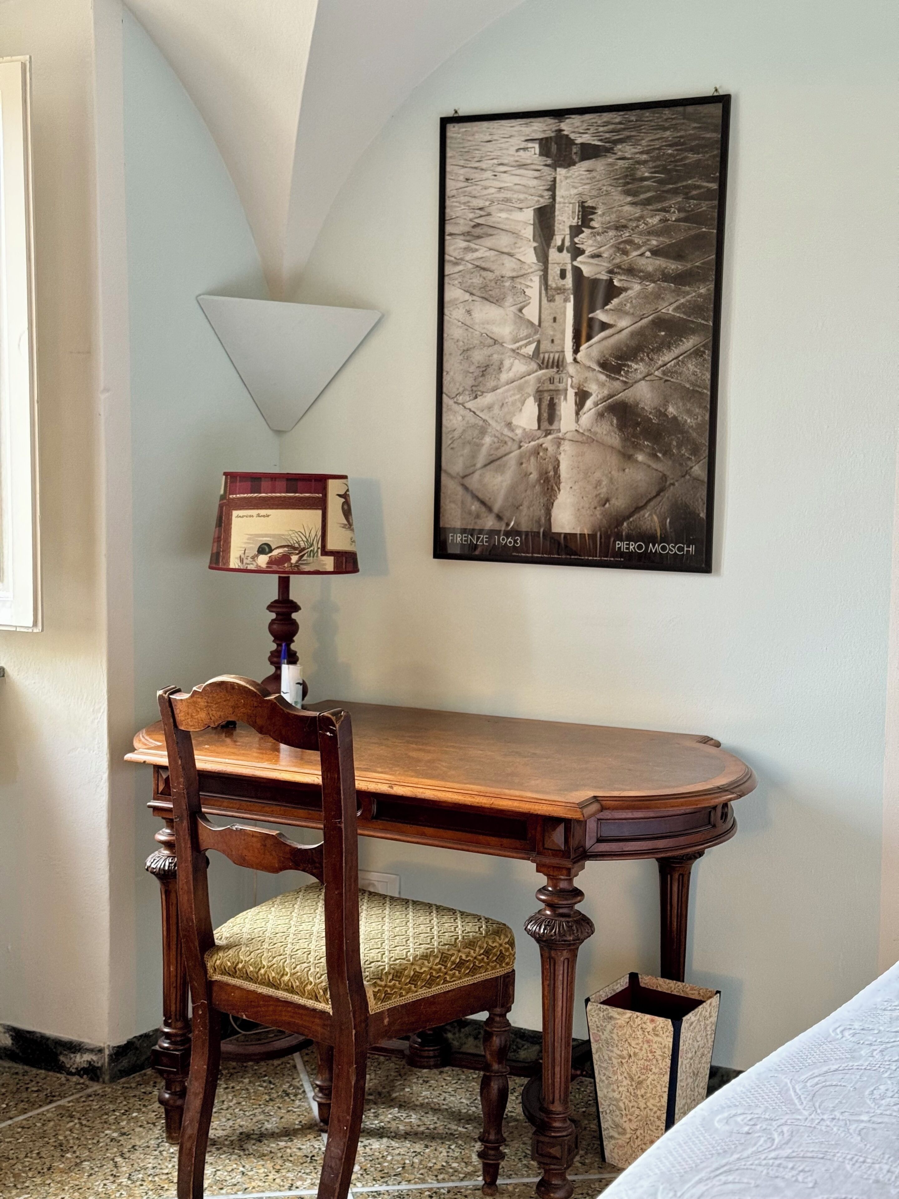Une pièce avec un bureau en bois, une chaise avec un siège en osier, un tableau encadré au mur et une lampe.
