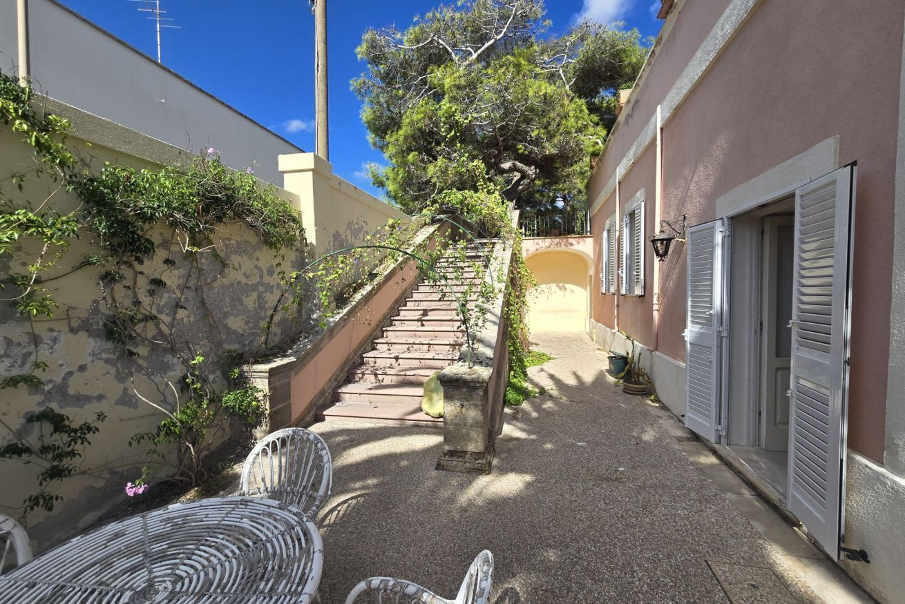 A narrow alleyway with a staircase leading to a building, surrounded by vegetation and a clear blue sky.