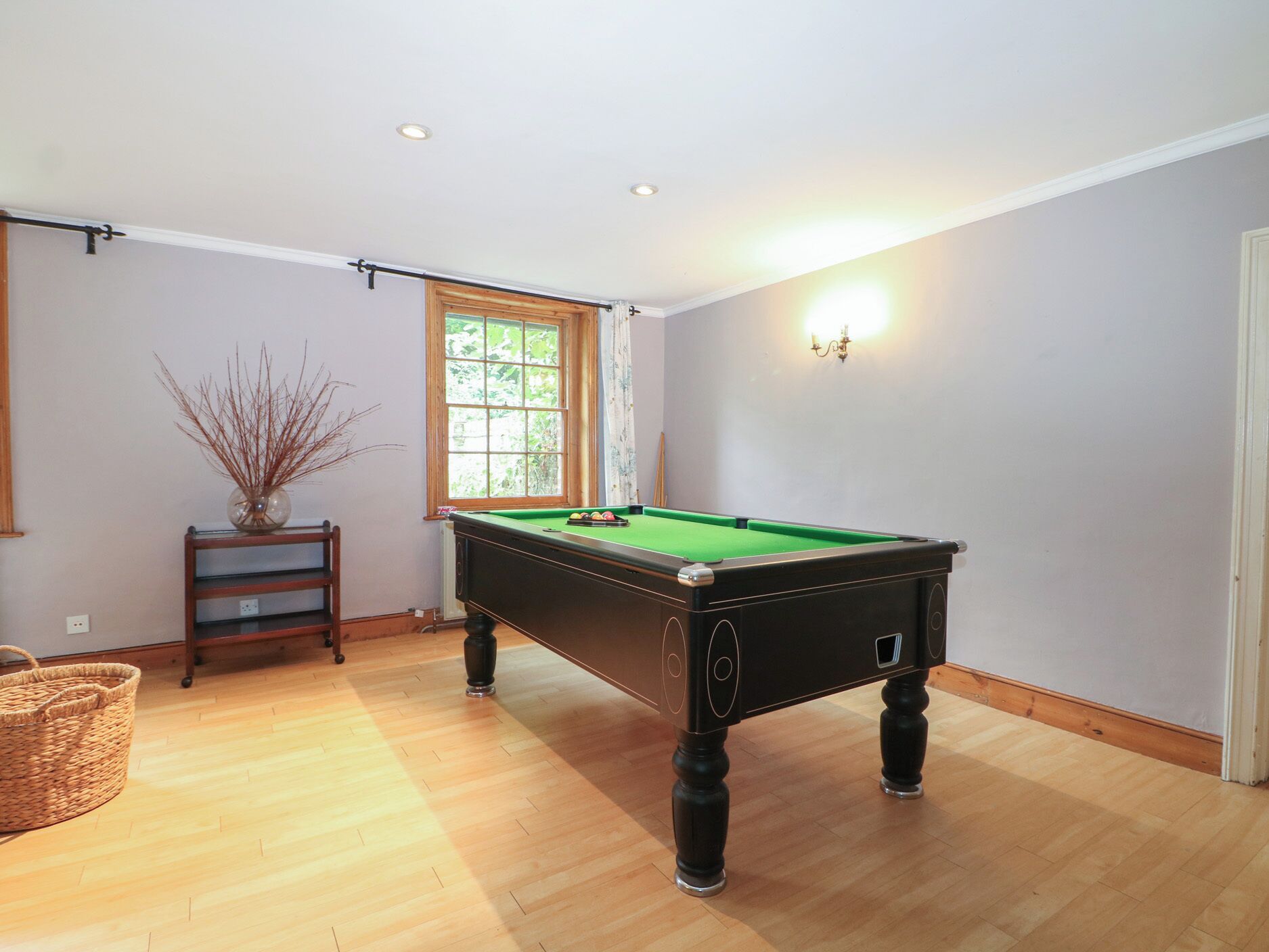 A pool table in a room with wooden flooring, a window with curtains, and a wall-mounted light fixture.