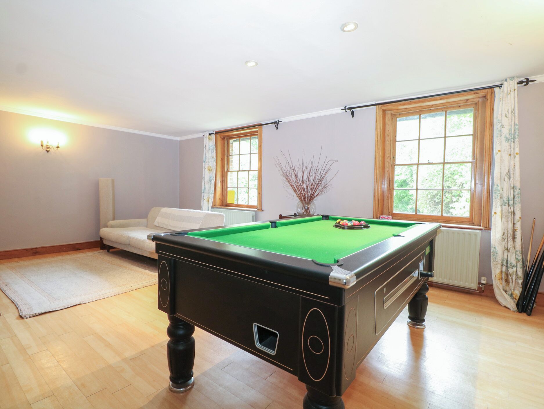 A pool table in a room with a white sofa, two windows with curtains, and a radiator.
