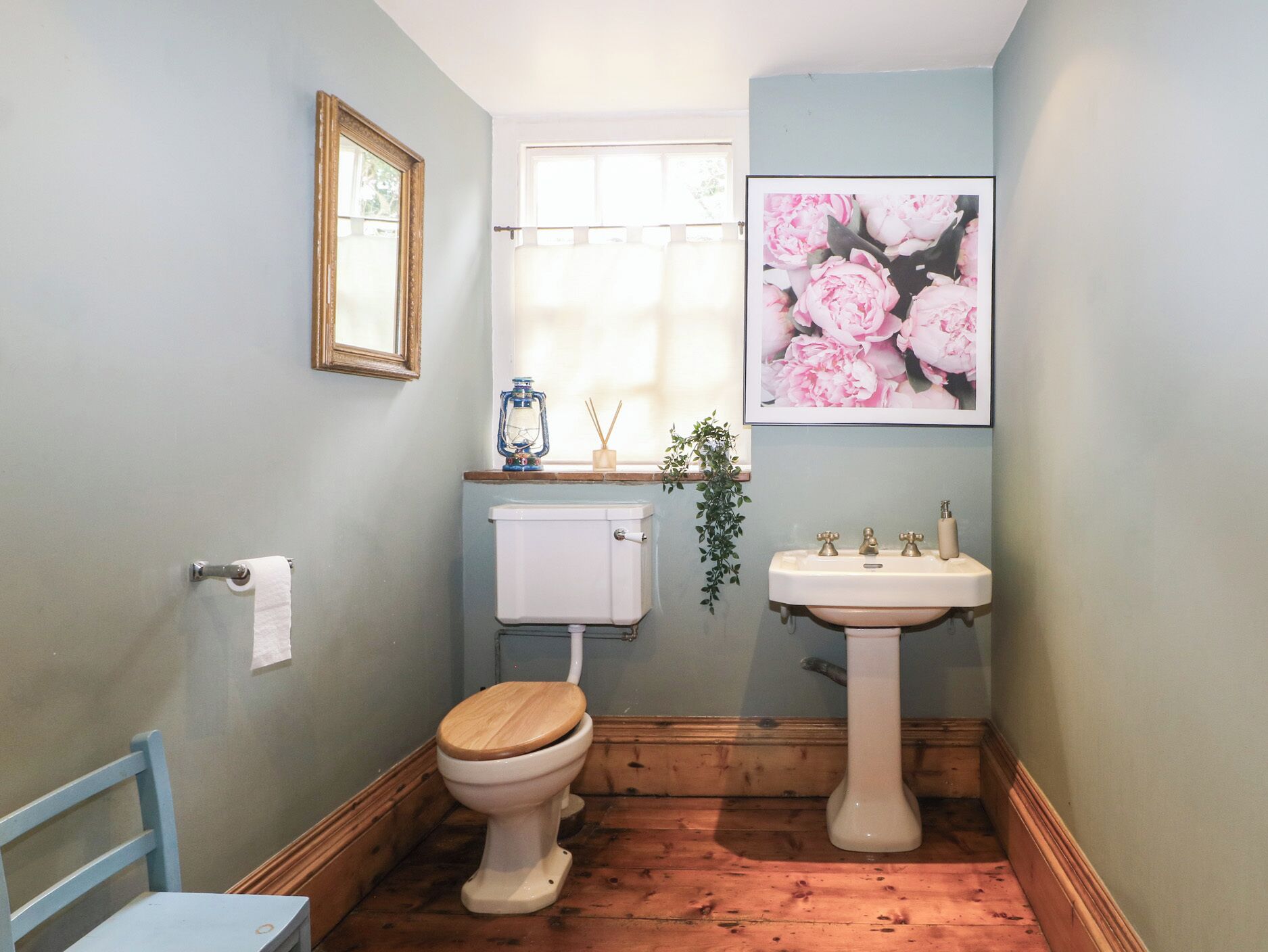 A bathroom with a wooden floor, a toilet with a wooden seat, a white sink, a framed picture of flowers, and a window with a curtain.