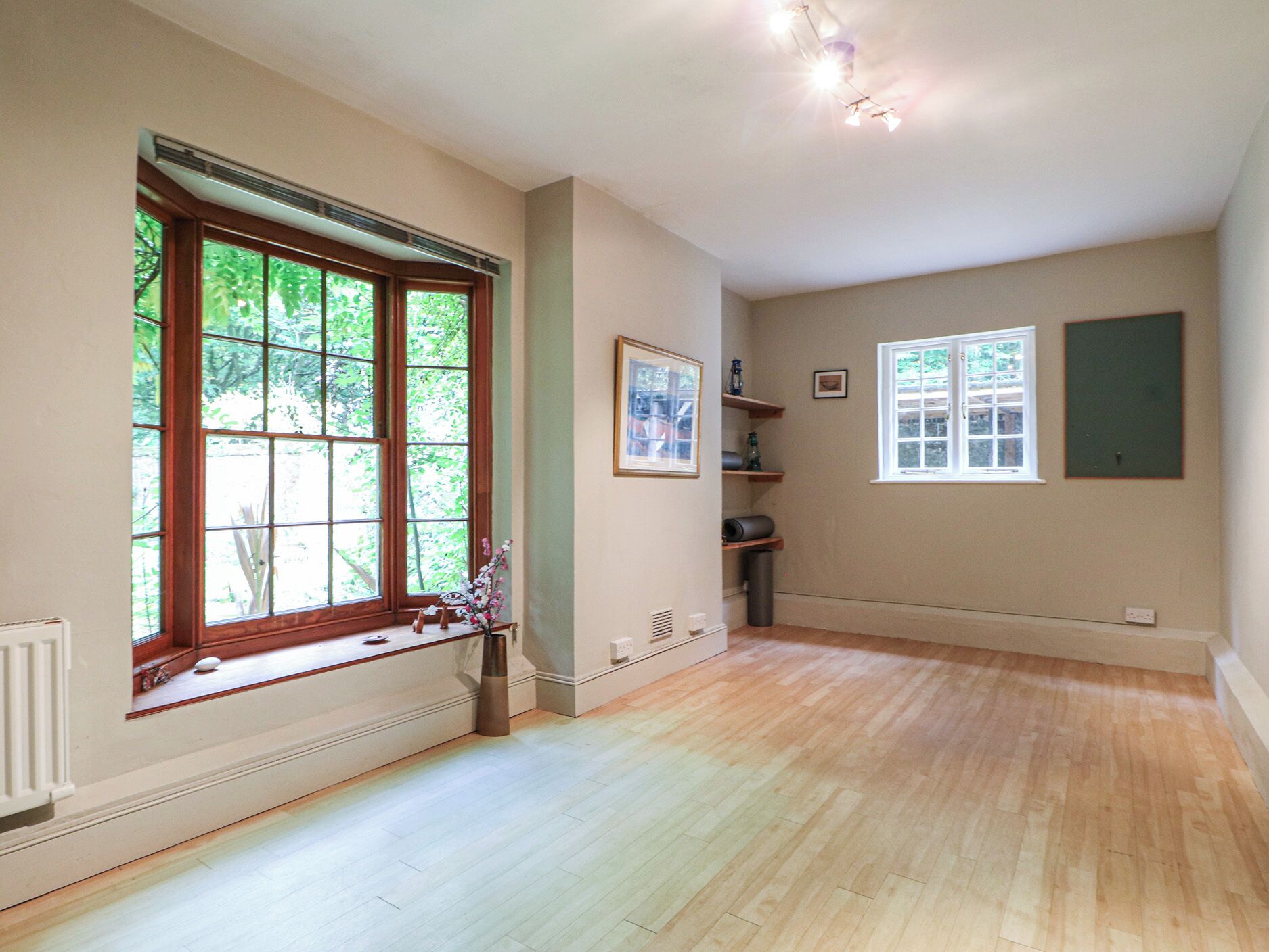 A spacious room with hardwood flooring, large windows with a view of greenery, built-in shelves, and a radiator.