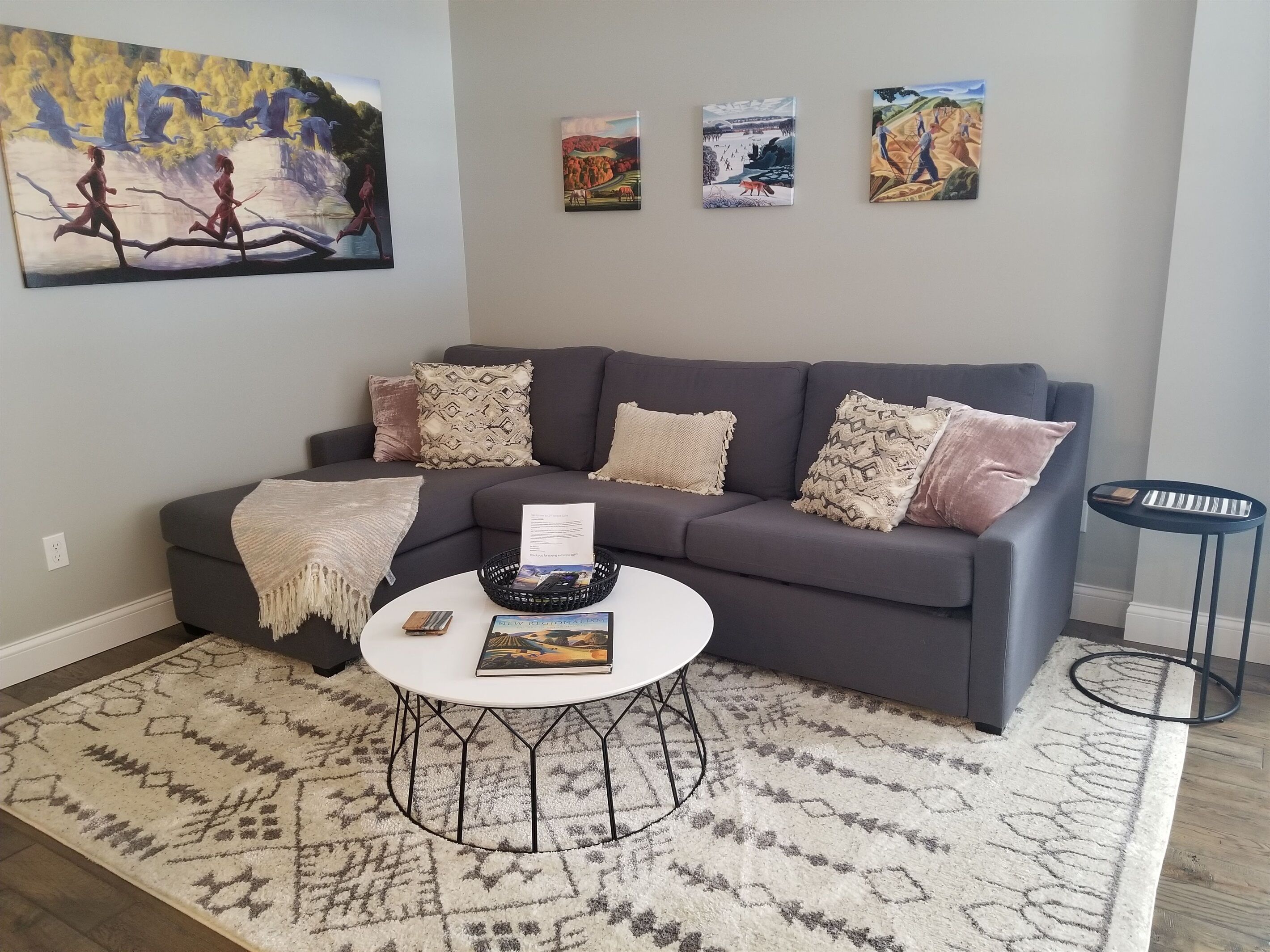 Un salon avec un canapé gris, des coussins décoratifs à motifs, une table basse blanche sur laquelle repose un magazine, et une petite table d’appoint noire.