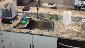 Een moderne keuken met een roestvrijstalen spoelbak, een koffiezetapparaat en een vaatwasser. Er is een eethoek met een tafel en stoelen, en een bank op de achtergrond.