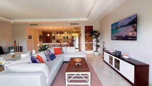 A modern living room with a white sectional sofa, a wooden coffee table, and a patterned rug. There is a kitchen area in the background with white cabinets and pendant lighting. - Tropical Garden Retreat 2BR in Plaza Puntarena (Río Hato)