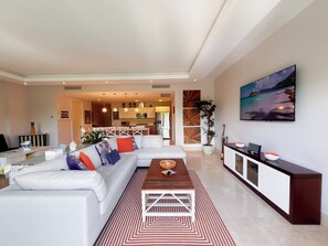 Ein modernes Wohnzimmer mit einem weißen Ecksofa, einem Holztisch und einem gemusterten Teppich. Im Hintergrund ist eine Küchenzeile mit weißen Schränken und Pendelleuchten zu sehen.