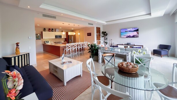A modern living room with a glass coffee table, white chairs, a sofa, and a TV mounted on the wall. - Tropical Garden Retreat 2BR in Plaza Puntarena (Río Hato)