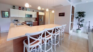 A modern kitchen with a central island, white bar stools, pendant lighting, and stainless steel appliances. - Tropical Garden Retreat 2BR in Plaza Puntarena (Río Hato)