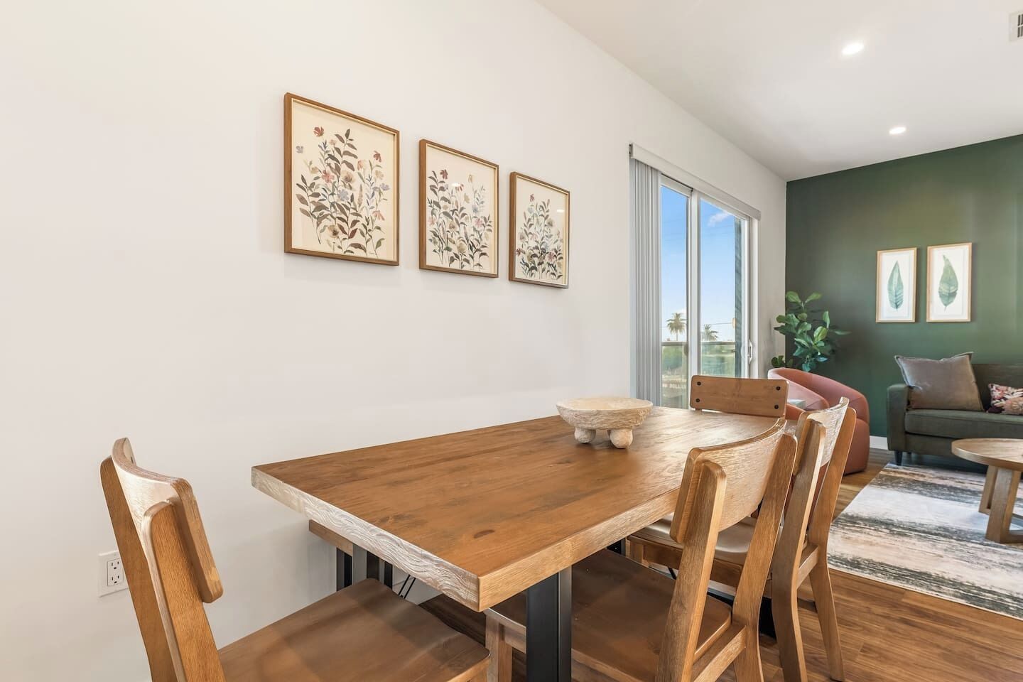 A dining area with a wooden table and chairs, framed botanical prints on the wall, and a view of the outdoors through a large window.
