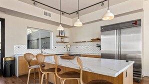 A modern kitchen with a marble island, wooden bar stools, a stainless steel refrigerator, and pendant lighting. - Light-Filled Home in K-Town | Parking, Gym & Views (Los Angeles)