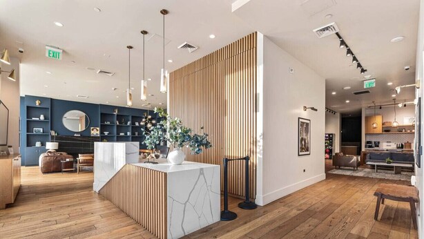 A modern hotel lobby with wooden flooring, a reception desk, a blue seating area, and a kitchen visible in the background. - Light-Filled Home in K-Town | Parking, Gym & Views (Los Angeles)