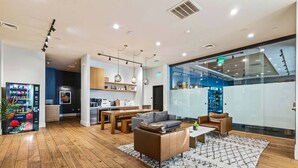 A modern hotel lobby with a wooden floor, a seating area with sofas and a coffee table, a kitchen in the background, and a glass wall with a bar area. - Light-Filled Home in K-Town | Parking, Gym & Views (Los Angeles)