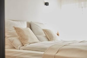 Ein Bett mit beiger Tagesdecke und Kissen, ein Nachttisch und eine Wandleuchte.