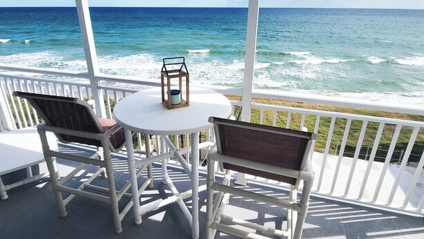 A balcony with white railings, two wicker chairs, a small round table, and a lantern. The view includes the ocean and a clear sky. - Salt Life Oasis - Direct Oceanfront, End Unit in Satellite Beach, FL (Satellite Beach)