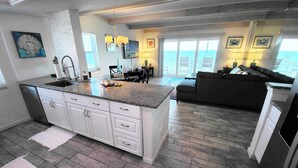 A modern kitchen with a large island, white cabinetry, and a grey countertop. There is a dining area with a table and chairs, and a dark-colored sofa in the living room. - Salt Life Oasis - Direct Oceanfront, End Unit in Satellite Beach, FL (Satellite Beach)