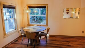 A dining area with a round table, four chairs, a window with a view of greenery, and a wall-mounted mirror. - Candler Cottage: Close 2 Everything "Asheville" (Candler)
