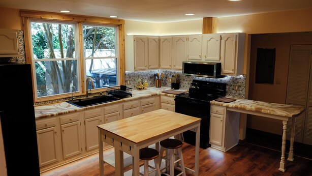 A kitchen with white cabinets, a wooden table, and a window overlooking a tree. - Candler Cottage: Close 2 Everything "Asheville" (Candler)