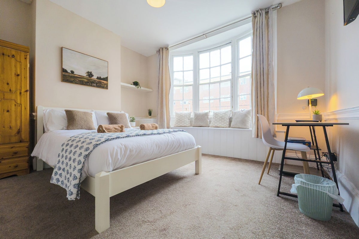 A bedroom with a bed, a desk, a chair, a window, and a plant.