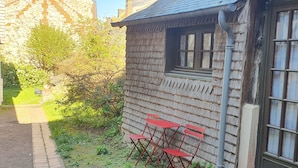 Ein kleines Holzgebäude mit Fenster und Tür, ein gepflasterter Weg und ein kleiner Garten mit einer Bank.