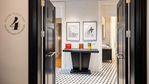 Un pasillo con suelo a cuadros blanco y negro, una puerta negra con un letrero con el número 4, y una mesa con dos cuadros enmarcados y una caja roja.