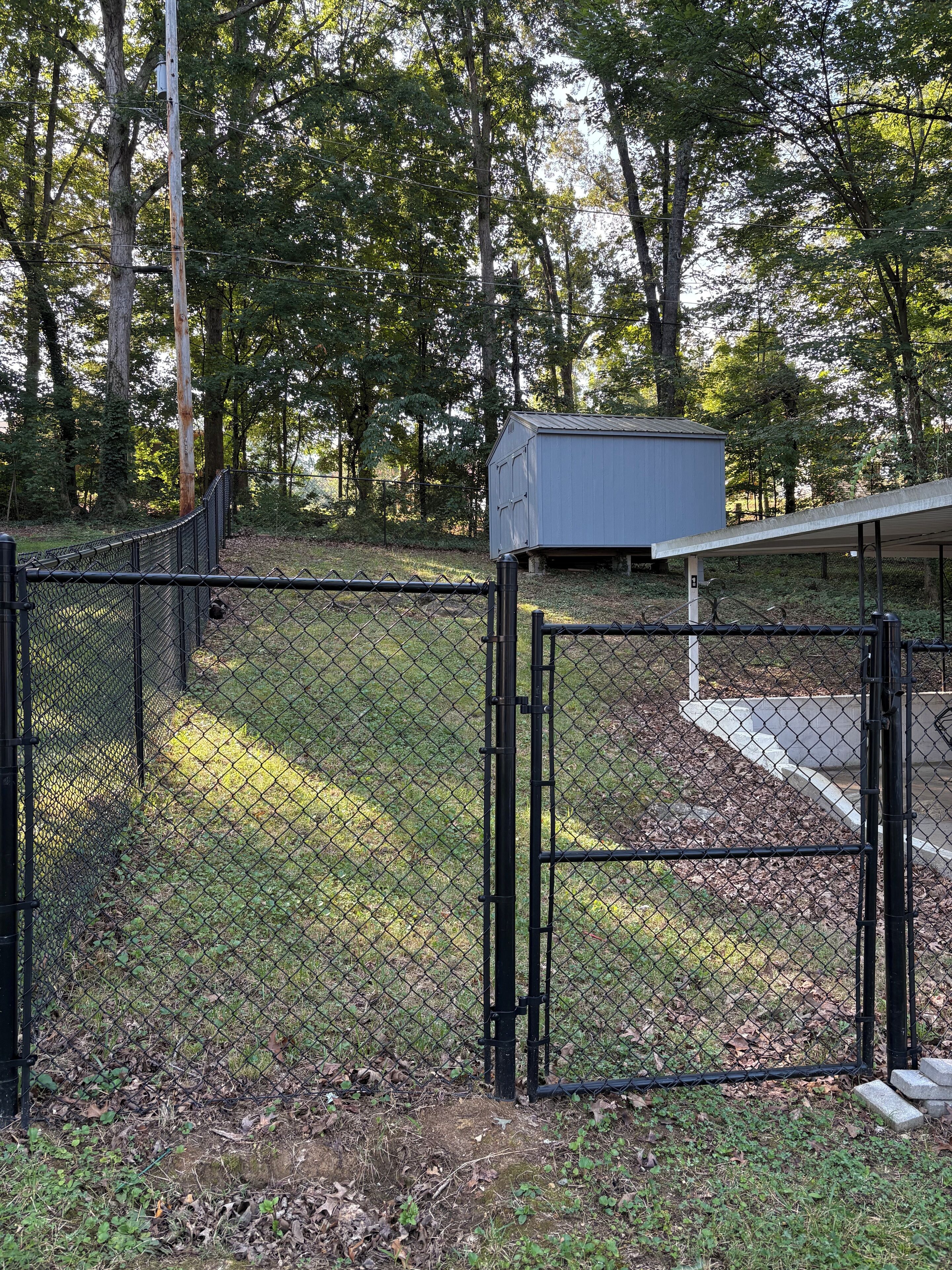 Un espace délimité par une clôture, avec un coffret électrique, une clôture en métal et des arbres en arrière-plan.