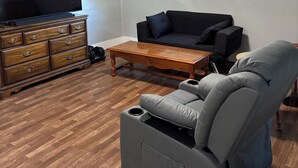 Ein Wohnzimmer mit einem Flachbildfernseher, einer Holzkommode, einem grauen Sofa und einem grauen Liegesessel.