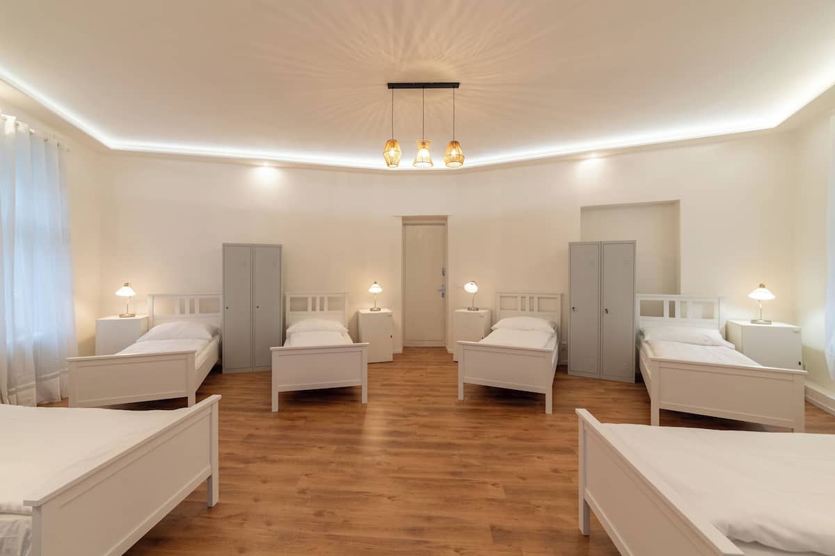 A spacious room with four white beds arranged in a square layout, wooden flooring, and a central ceiling light fixture.