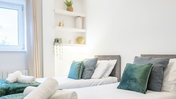 A hotel room with two beds, white linens, green and blue pillows, a shelf with decorative items, and a window with curtains. - Modern 2BR Apartment with Water Views (Cardiff)