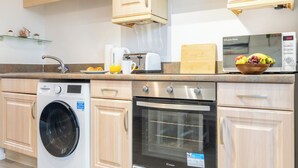 A compact kitchen with a washing machine, oven, microwave, and a fruit bowl on the counter. - Modern 2BR Apartment with Water Views (Cardiff)