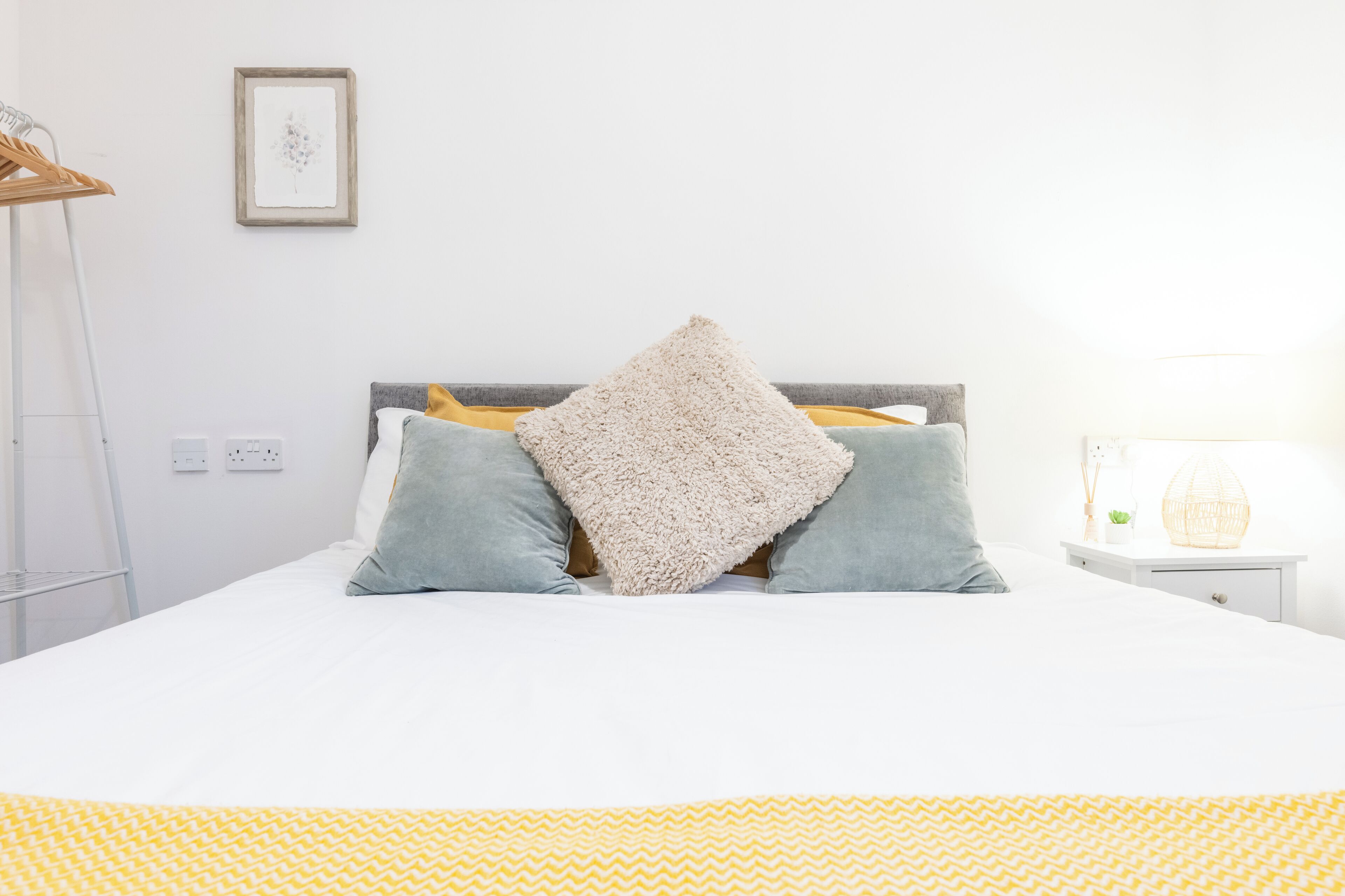 Uma cama bem arrumada com edredom branco e amarelo estampado, duas almofadas cinzas e um cobertor bege felpudo.