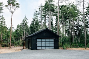 Uma pequena cabana de madeira escura com garagem, rodeada por altas árvores de pinheiro.