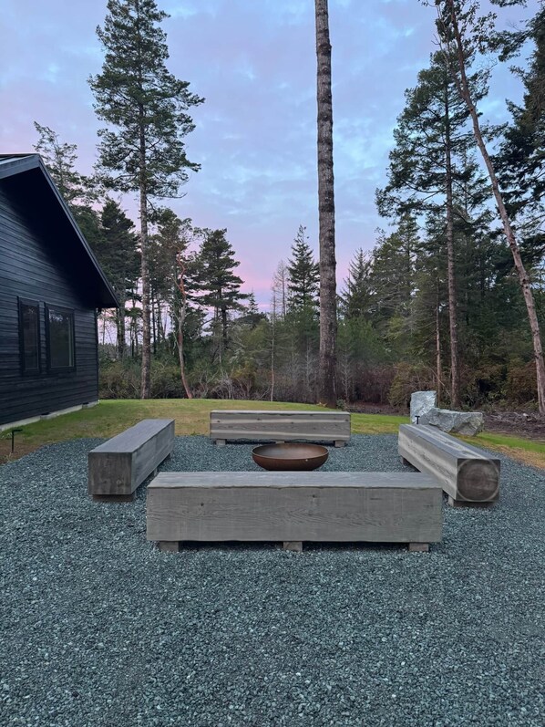 A rustic outdoor seating area with benches and a fire pit, surrounded by tall trees and a cabin. - Bandon Heath - Walk to Bandon Dunes Golf Resort (Bandon)