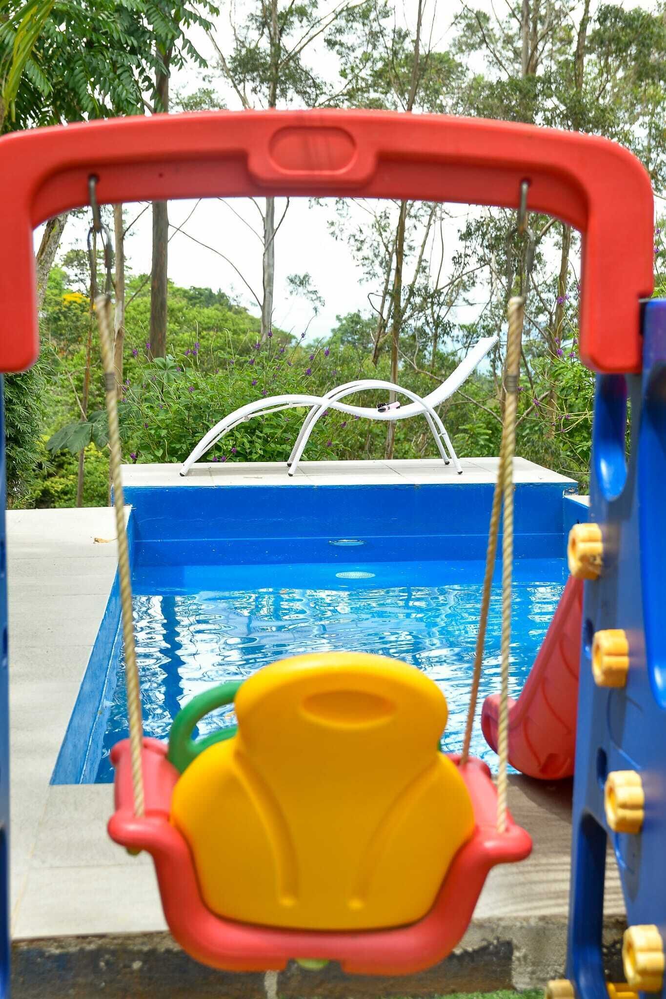 A children's swing set over a pool with a view of trees and greenery.