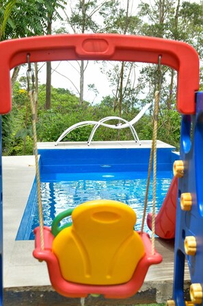 Une balançoire pour enfants installée au-dessus d’une piscine, avec vue sur les arbres et la verdure.