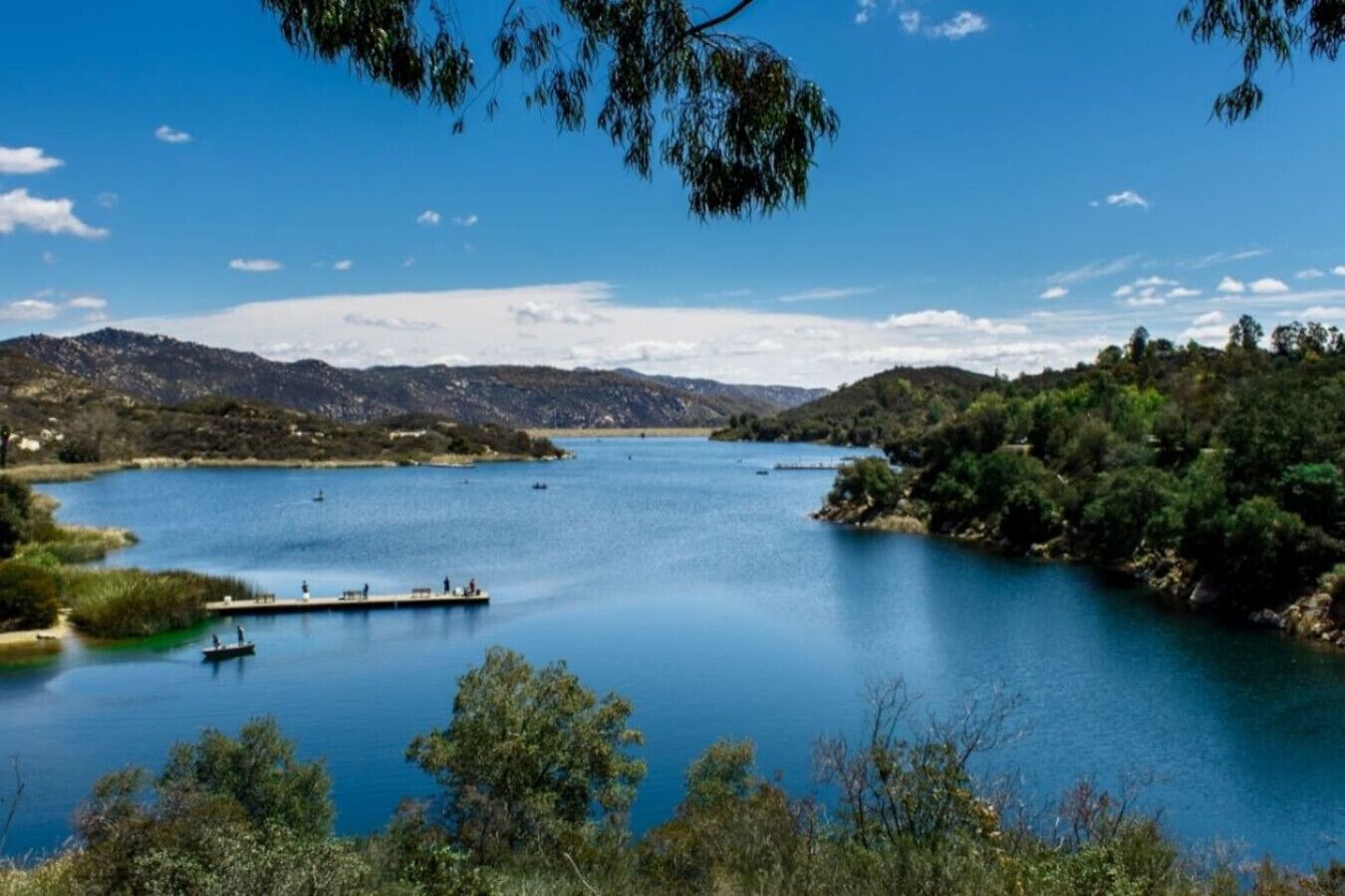 Un lago tranquilo con un muelle y barcos, rodeado de colinas y árboles.