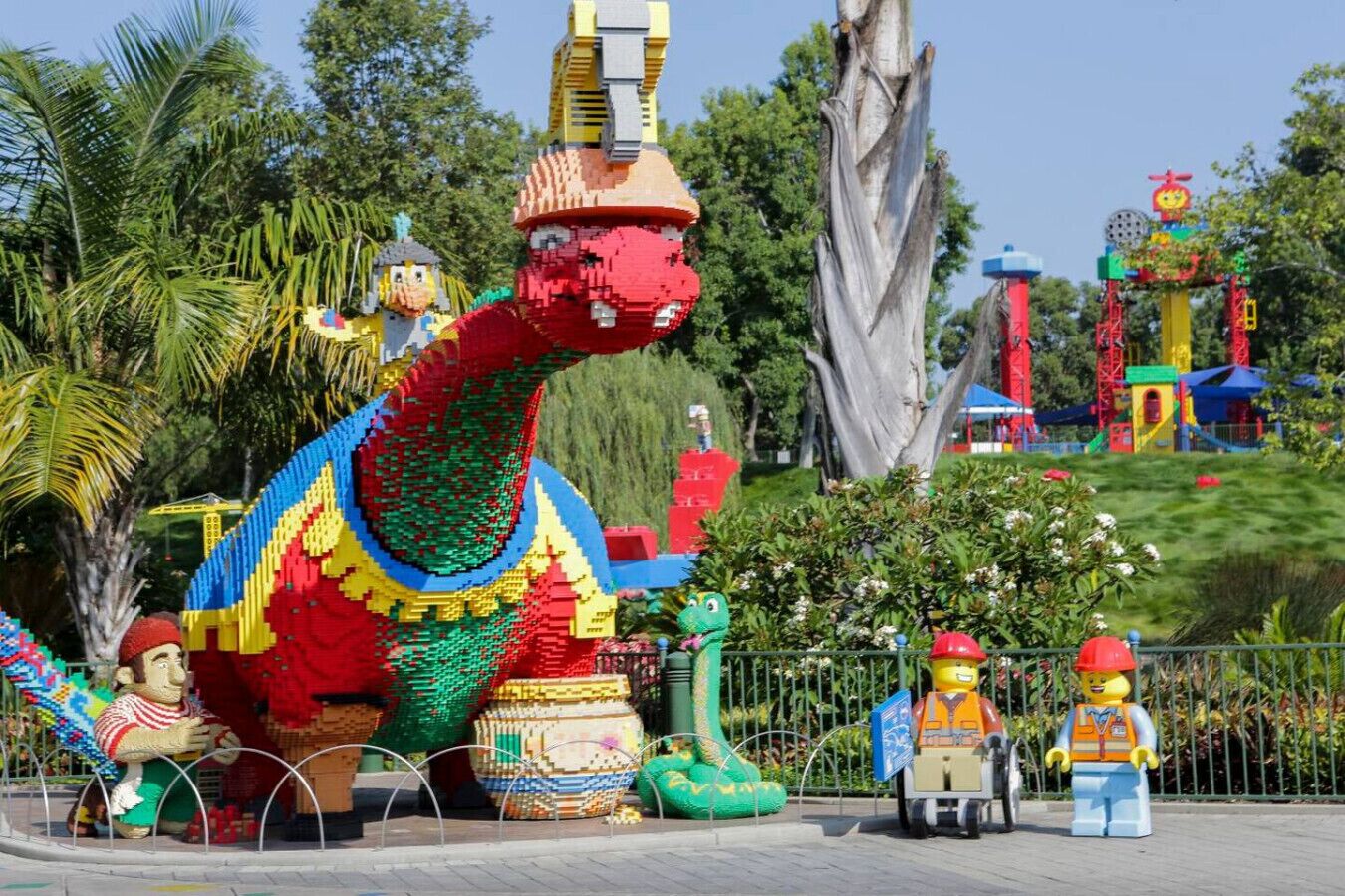 Eine farbenfrohe Dinosaurier-Statue, umgeben von kleineren Figuren und einem Spielplatz im Hintergrund.