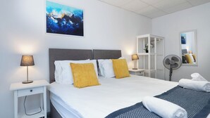Habitación de hotel con cama, lámparas de noche, un ventilador, un espejo y un cuadro en la pared.
