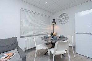 Ein gemütlicher Raum mit einem runden Esstisch, weißen Stühlen, einem grauen Sofa, einem Heizkörper, einer Wanduhr und einer weißen Tür.