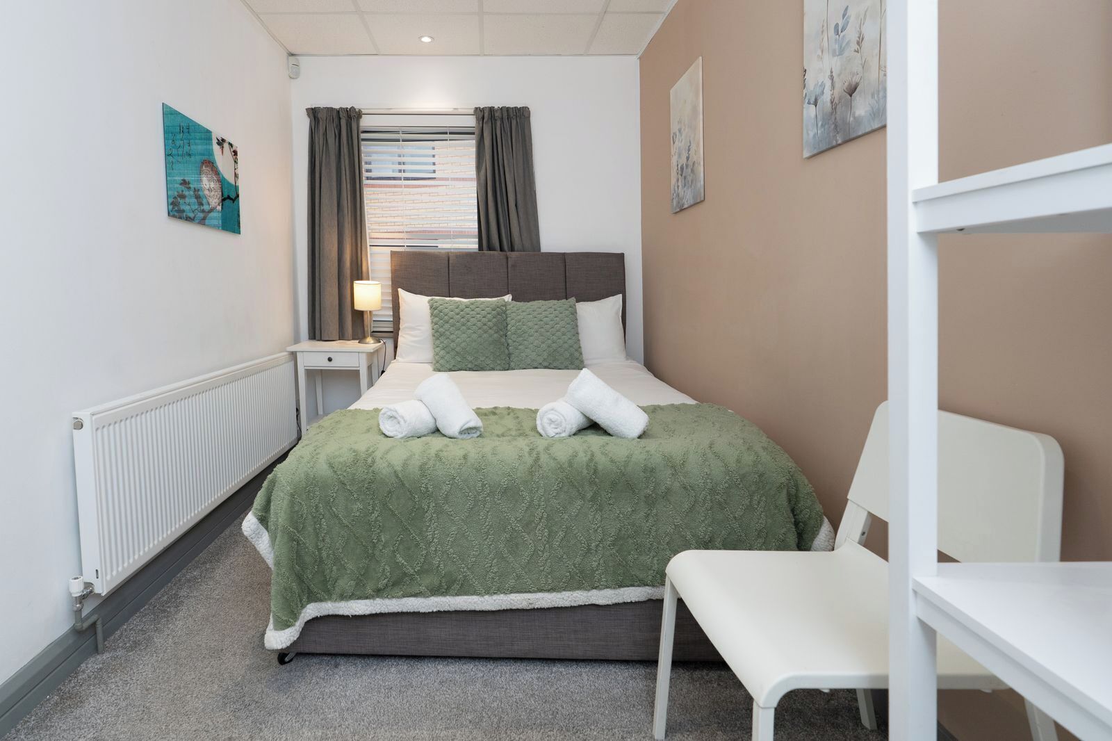 Ein Hotelzimmer mit Bett, Sessel, Heizkörper, einem Fenster mit Vorhängen und Bildern an den Wänden.