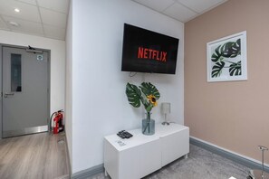 Ein Empfangsbereich mit einem Wandfernseher, der 'NETFLIX' zeigt, einer weißen Konsole mit einer Vase und einer Pflanze sowie einer gerahmten Pflanzenkunst am Wand.