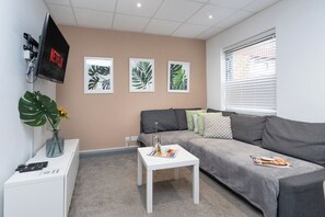 Ein Wohnzimmer mit grauem Sofa, einem weißen Couchtisch, einem an der Wand montierten Fernseher und drei gerahmten Pflanzenbildern.