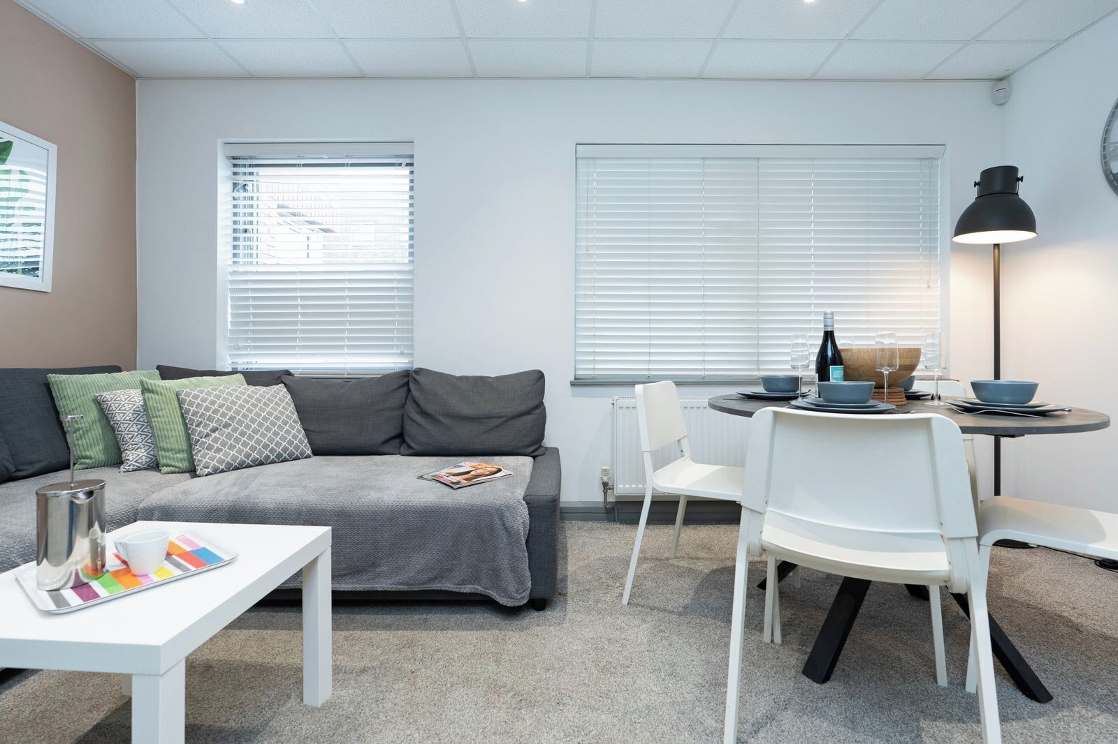 Ein modernes Wohnzimmer mit einem grauen Sofa, weißen Stühlen, einem kleinen Esstisch und einer Lampe.