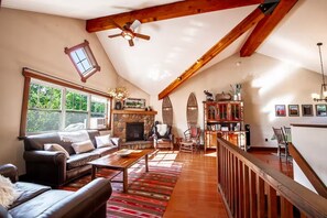 Una sala de estar espaciosa con chimenea, techo alto con vigas a la vista, un ventanal grande y una zona de estar acogedora.