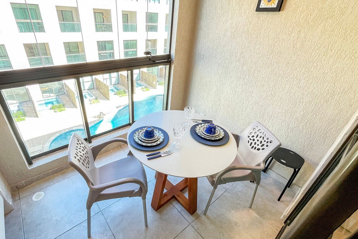 Un coin repas avec une table et des chaises, une fenêtre donnant sur un bâtiment et une piscine, et un tableau encadré accroché au mur.