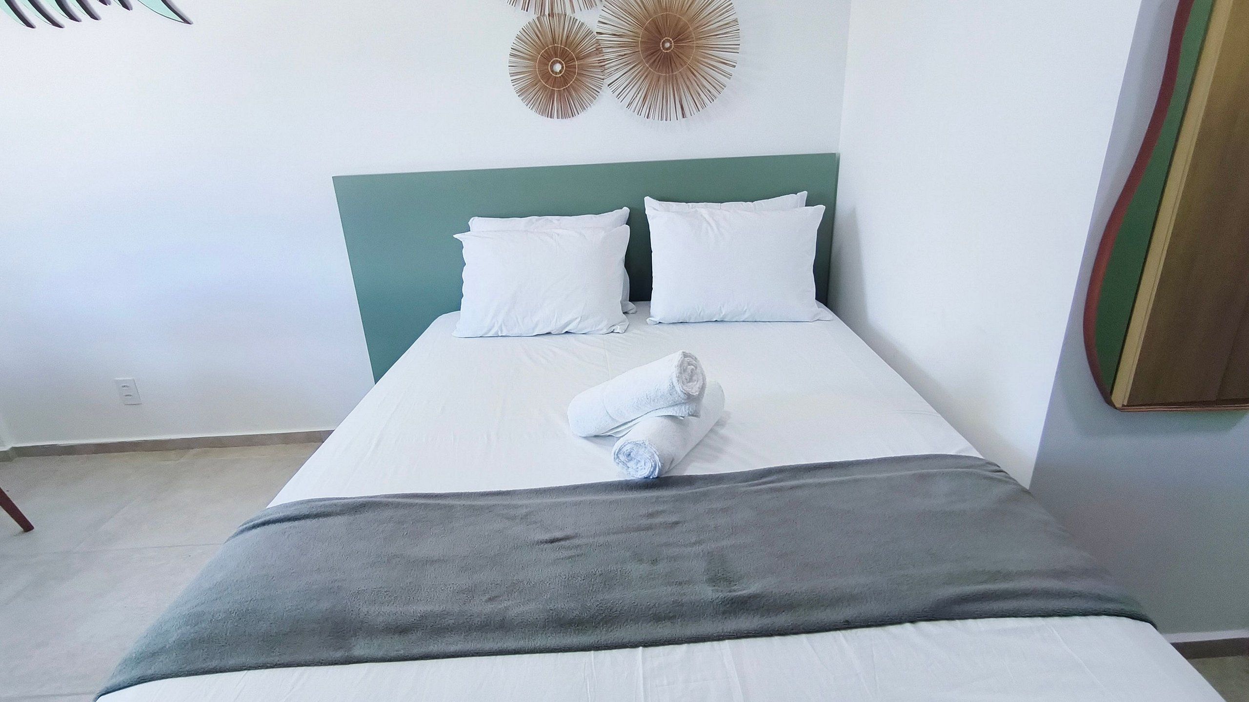 A bed with white linens, a grey blanket, and a rolled towel.