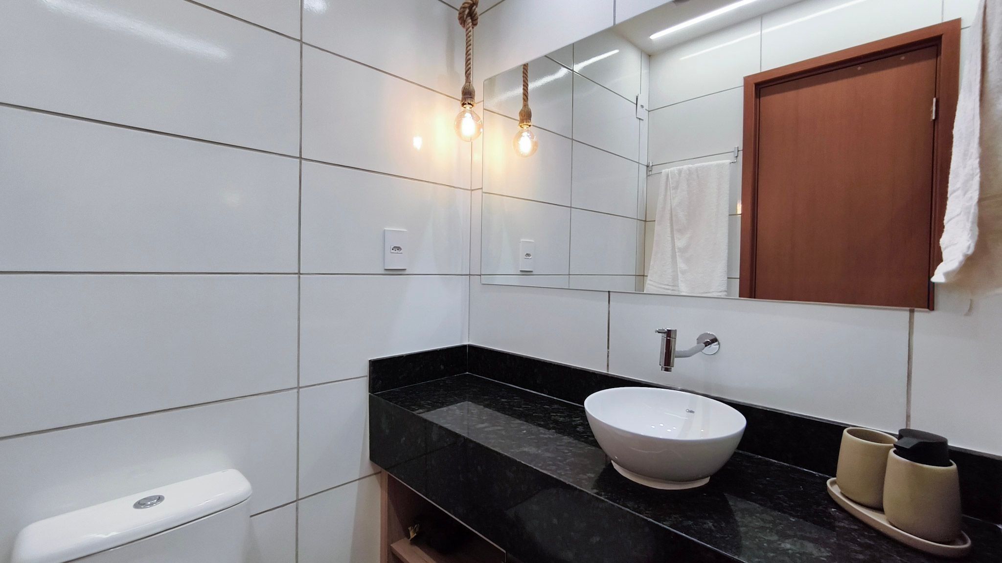 Un baño moderno con baldosas blancas, encimera de mármol negro, lavabo blanco y puerta de madera.