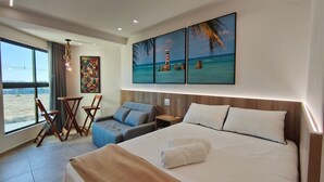 Una camera d'albergo con un letto, un'area salotto con sedie, un tavolino, un quadro raffigurante una spiaggia e vista sull'esterno.