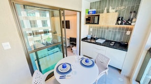 Een kleine keuken met een magnetron, broodrooster en koffiezetapparaat. Er staat een eettafel met blauwe en witte borden en een stoel.