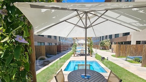 A poolside area with a table and chairs, a large umbrella, and a view of a pool and building in the background.